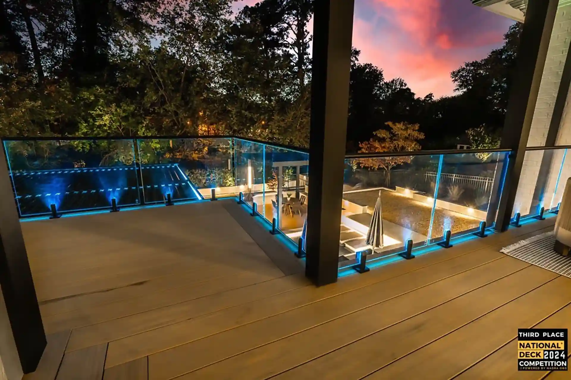 Photo of a modern deck with glass railings, blue LED lighting, and a scenic sunset view.